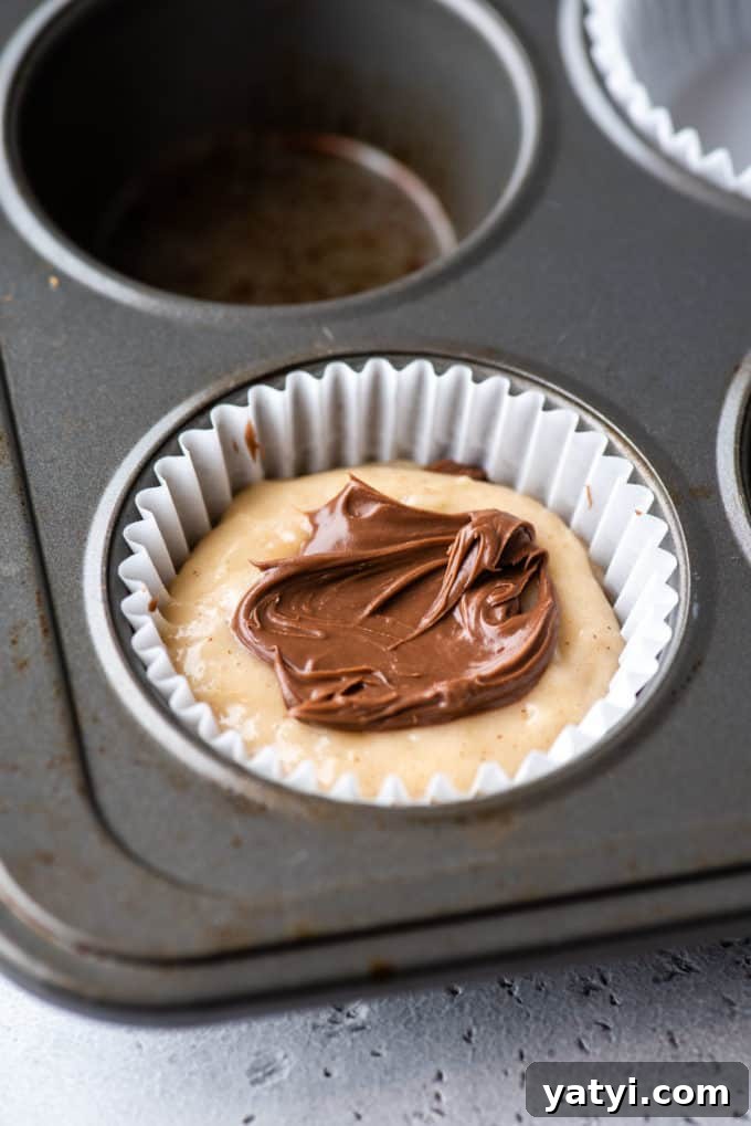Nutella Swirl Muffins Recipe: Moist Chocolate-Hazelnut Muffins 9 a muffin pan with a white muffin liner in the corner spot and some muffin batter in the bottom of it and layers of muffin batter and nutella inside the liner