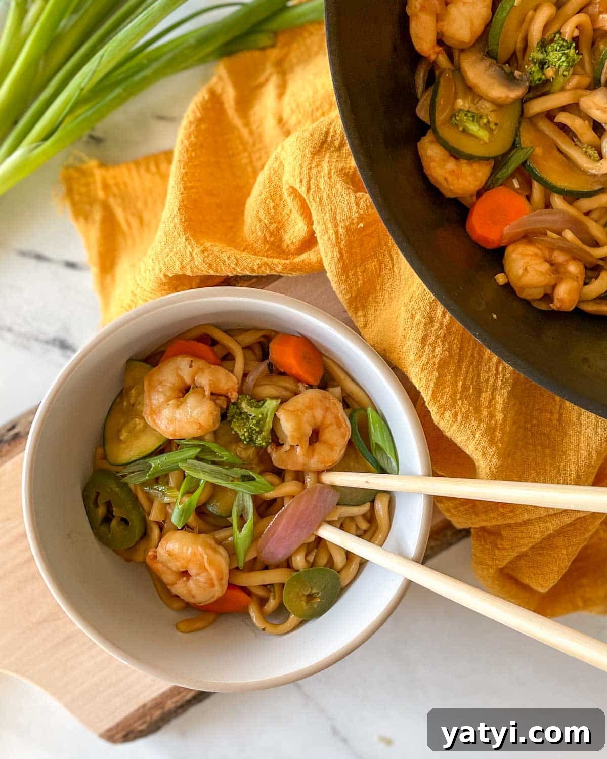 A white bowl of Shrimp Yaki Udon sits on a rustic wooden cutting board with chopsticks with a yellow linen and green onions.