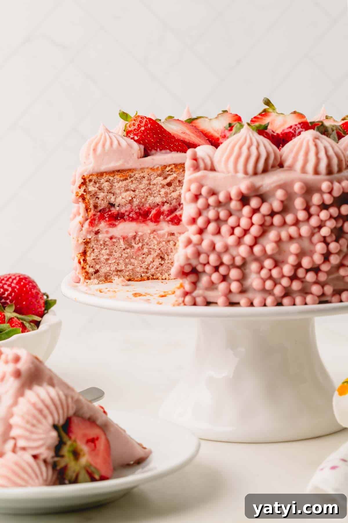 Moist Homemade Strawberry Layer Cake Recipe 2 A two layer frosted strawberry cake on a cake stand with a slice missing.