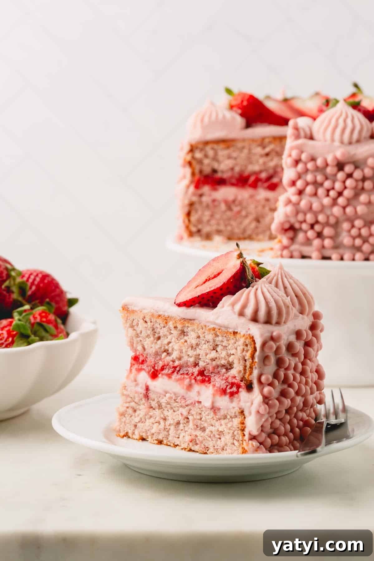 Moist Homemade Strawberry Layer Cake Recipe 3 A slice of layered frosted strawberry cake on a plate next to the rest of the cake on the stand.