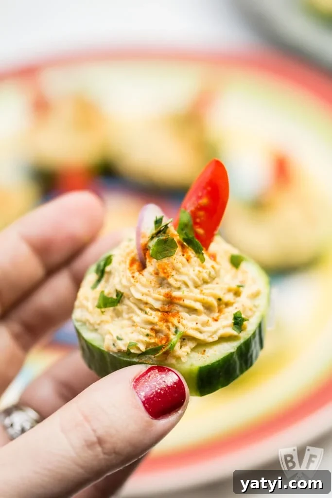 Layered Taco Dip with Refried Beans for Parties and Game Day 17 Close up of a hand holding a cucumber slice topped with a spiced chickpea purΓ©e.