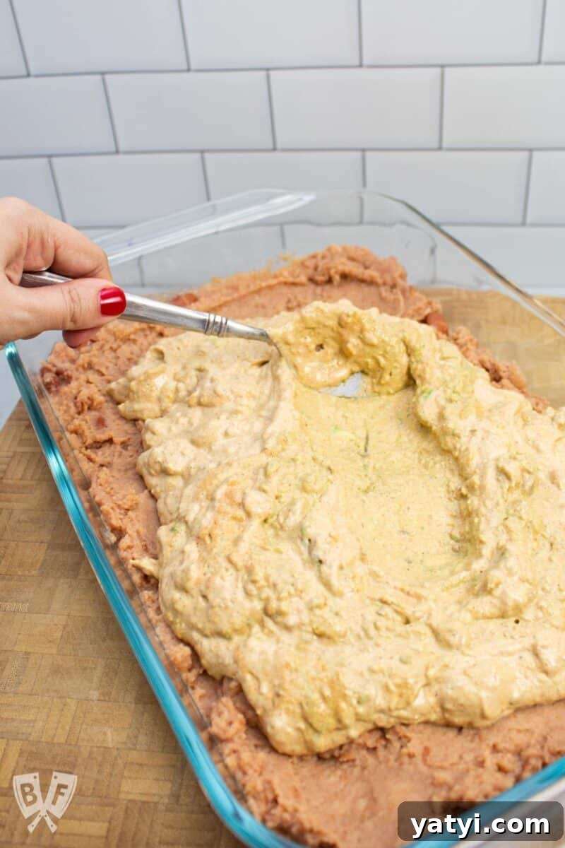 Layered Taco Dip with Refried Beans for Parties and Game Day 6 Smoothing a seasoned avocado mixture over refried beans in a serving dish for layered taco dip.