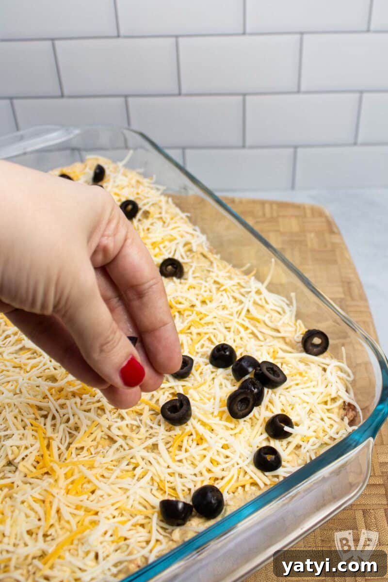 Layered Taco Dip with Refried Beans for Parties and Game Day 8 Sprinkling black olives on top of a layered taco dip.