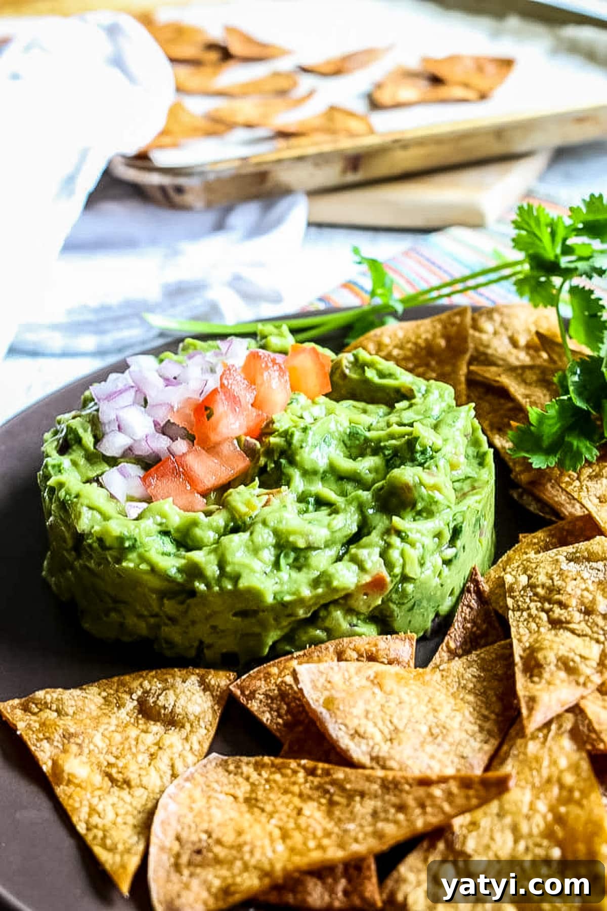 Crispy Oven-Baked Tortilla Chips Recipe You Can Make at Home 4 tortilla chips laying on a brow platter next to guacamole, in the back a baking sheet has freshly made chips.