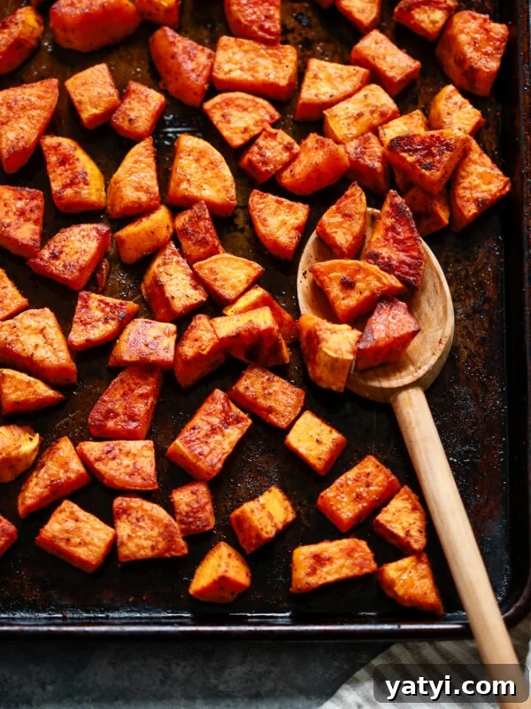 Crispy Air Fryer Sweet Potatoes: Quick Roast in 20 Minutes 10 Chunks of roasted sweet potato on a dark baking sheet, some scooped up with a wooden spoon. The potatoes are golden brown and seasoned, with a striped kitchen towel partially visible in the corner.