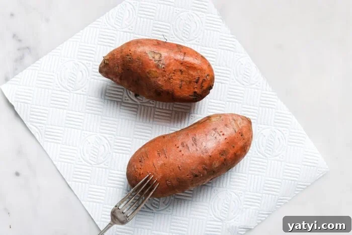 Crispy Air Fryer Sweet Potatoes: Quick Roast in 20 Minutes 4 Two whole Air Fryer Baked Sweet Potatoes rest on a white textured paper towel on a marble surface, with a fork placed beside them.