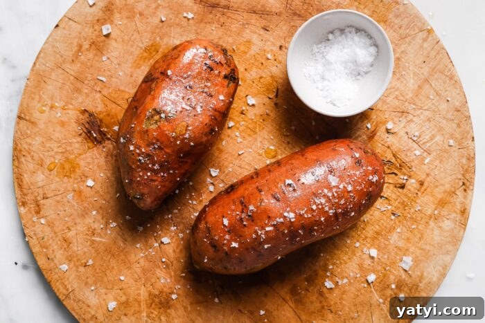 Crispy Air Fryer Sweet Potatoes: Quick Roast in 20 Minutes 5 Two Air Fryer Baked Sweet Potatoes rest on a round wooden cutting board, sprinkled with coarse sea salt. A small white bowl filled with sea salt sits beside the sweet potatoes.