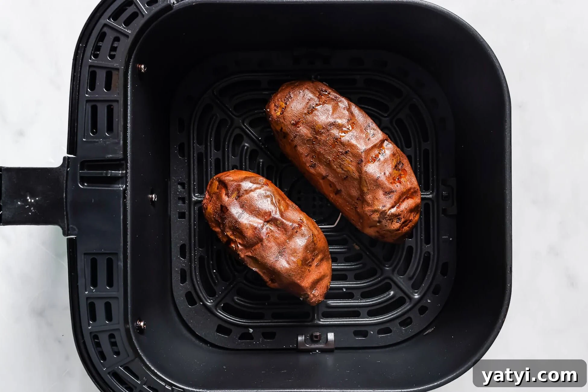 Crispy Air Fryer Sweet Potatoes: Quick Roast in 20 Minutes 7 Two cooked Air Fryer Baked Sweet Potatoes sit inside the basket of an air fryer on a white marble surface.