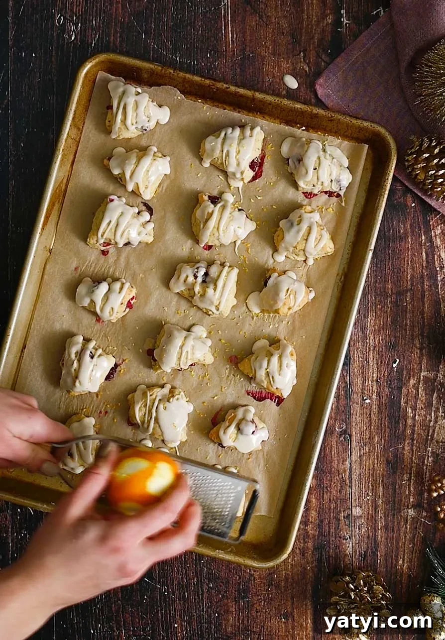 Vegan Holiday Afternoon Tea Menu & Festive Recipes 5 Someone zesting an orange over small, glazed cranberry scones on a baking sheet