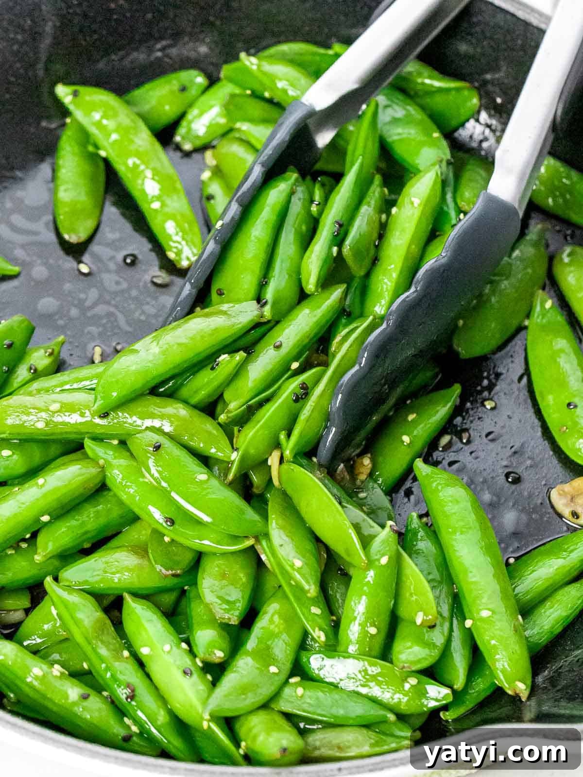 Garlic sesame sugar snap peas stir fried in a dark pan with a pair of tongs.