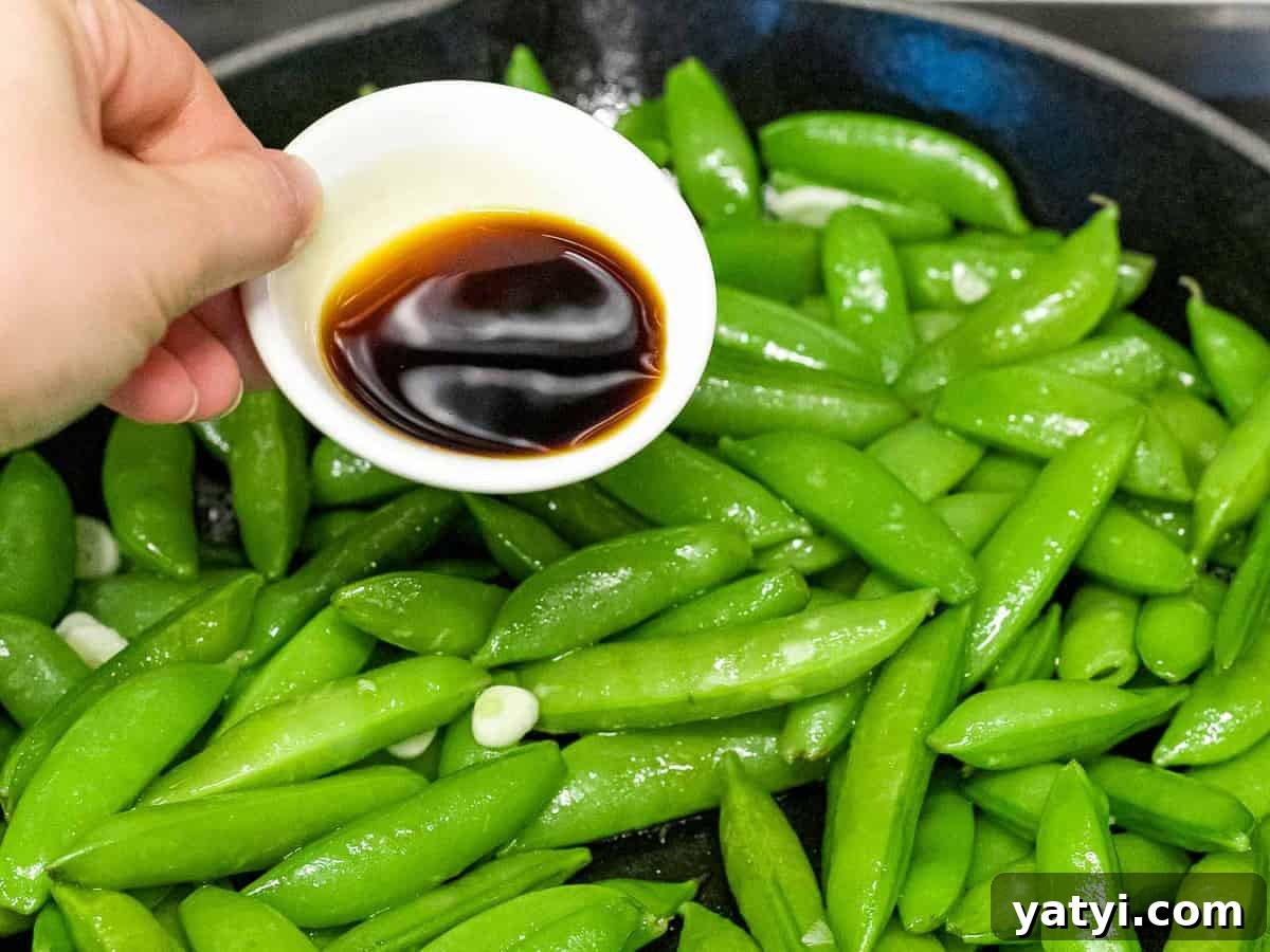 Soy sauce being added to sugar snap peas sauteed in a pan.