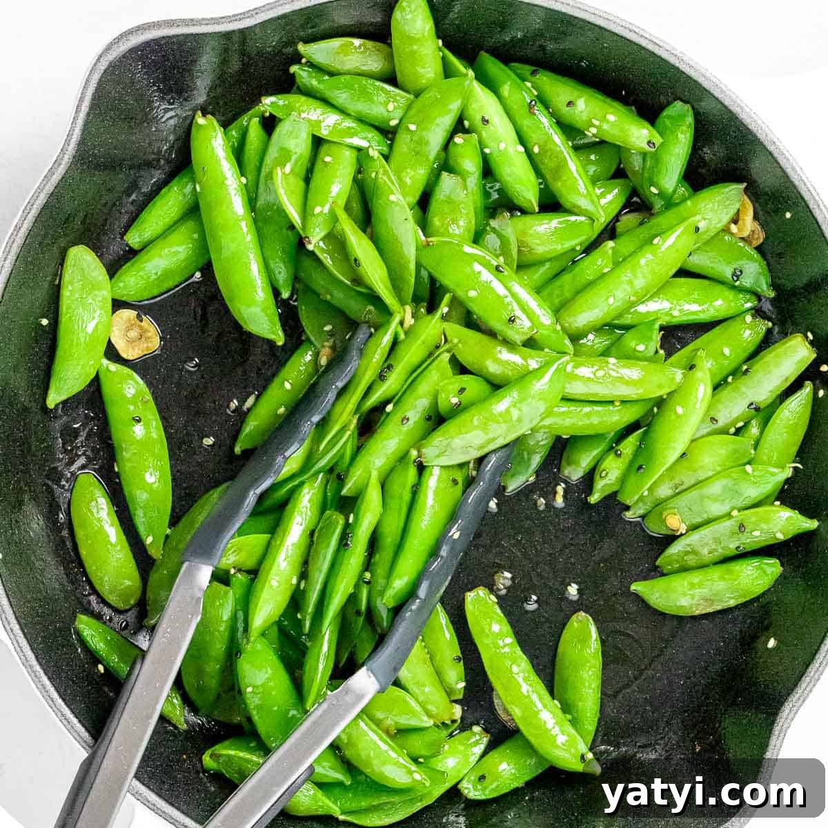 Stir fried sugar snap peas with garlic, black and white sesame seeds, and soy sauce in a dark pan with tongs.