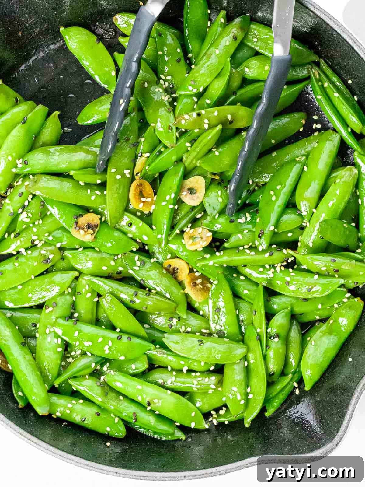 Stir fried sesame sugar snap peas with slices of garlic in a pan with a pair of tongs.