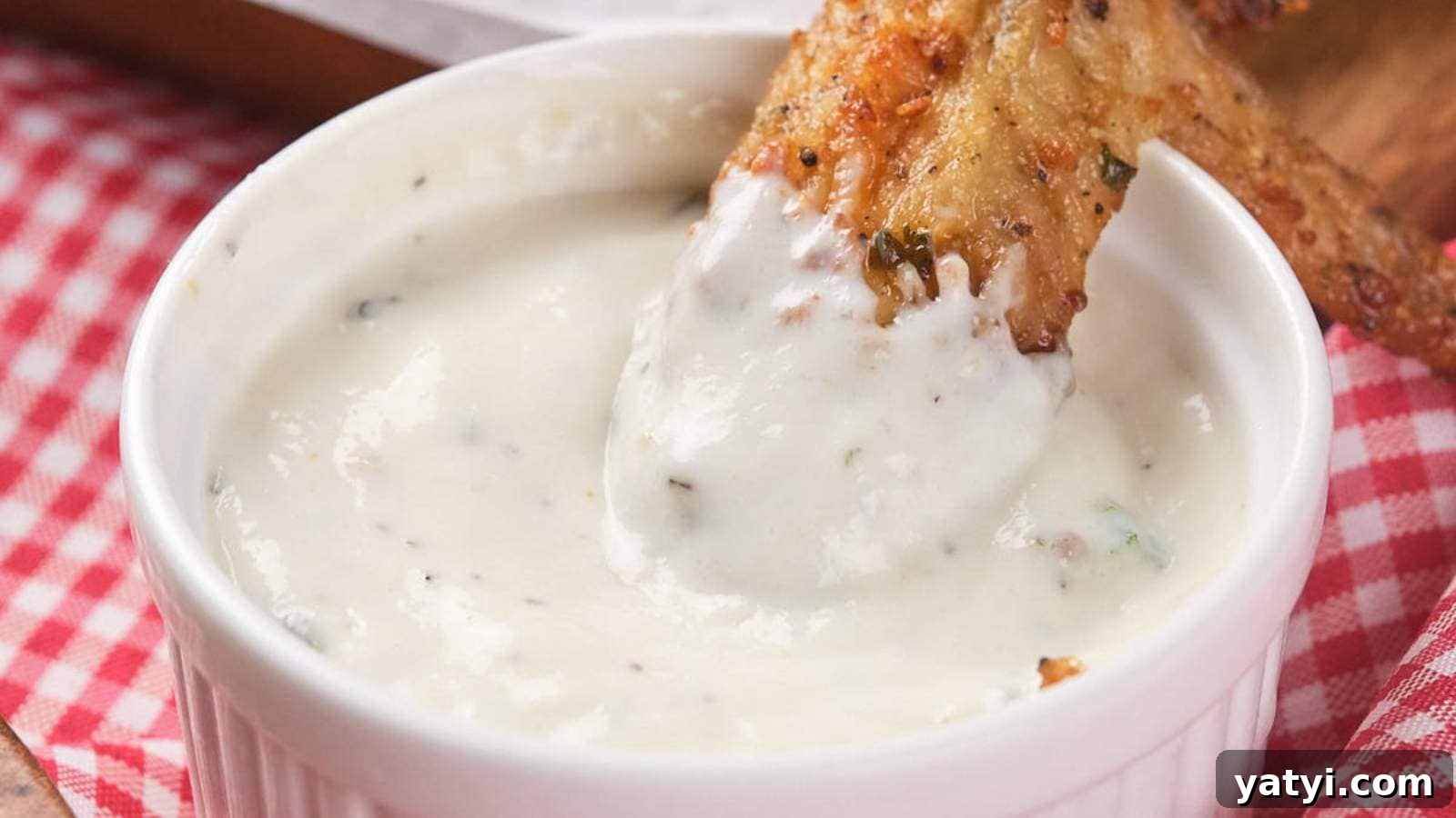 21 Snack Swaps That Beat Junk Food Without Sacrificing Flavor 16 Chicken wing dipped in a ranch sauce in a small white bowl, placed on a red and white checkered cloth.