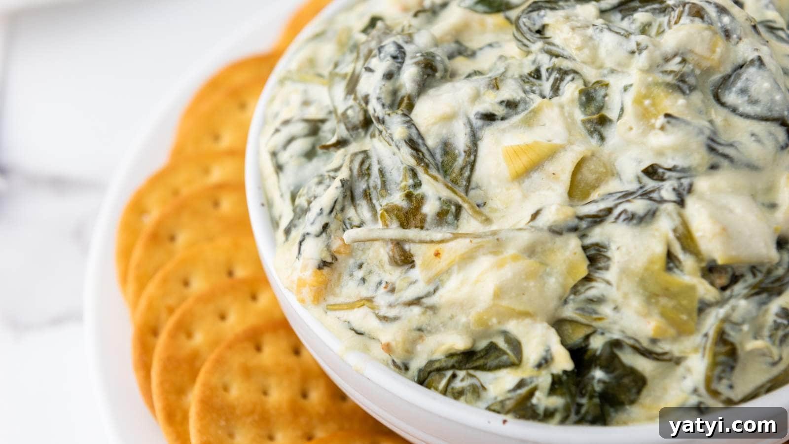 21 Snack Swaps That Beat Junk Food Without Sacrificing Flavor 7 A bowl of creamy spinach artichoke dip with a side of round crackers on a white surface.