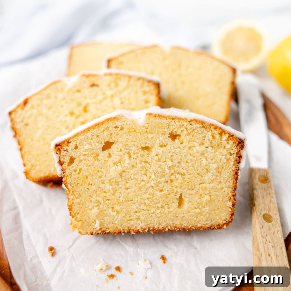 Copycat Starbucks Lemon Loaf Recipe: Moist Citrus Pound Cake Tutorial 2 Sliced lemon loaf cake with a light glaze on top, arranged on parchment paper next to a wooden-handled knife.