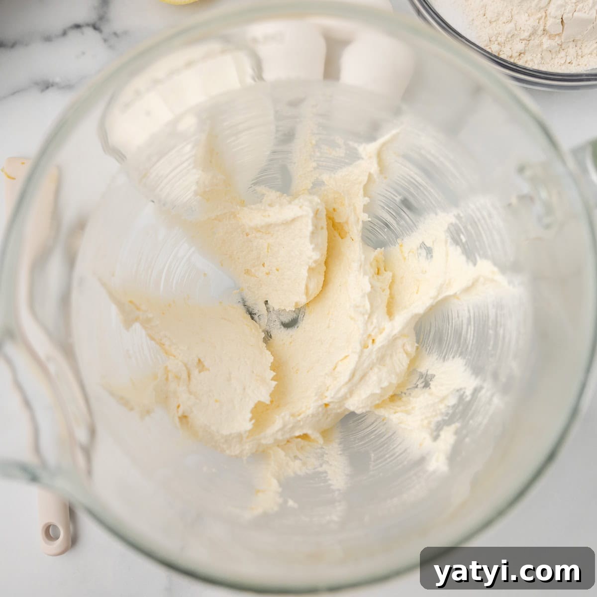 Copycat Starbucks Lemon Loaf Recipe: Moist Citrus Pound Cake Tutorial 4 A glass mixing bowl containing creamed butter and sugar, with a spatula and a bowl of flour visible nearby on a marble countertop.