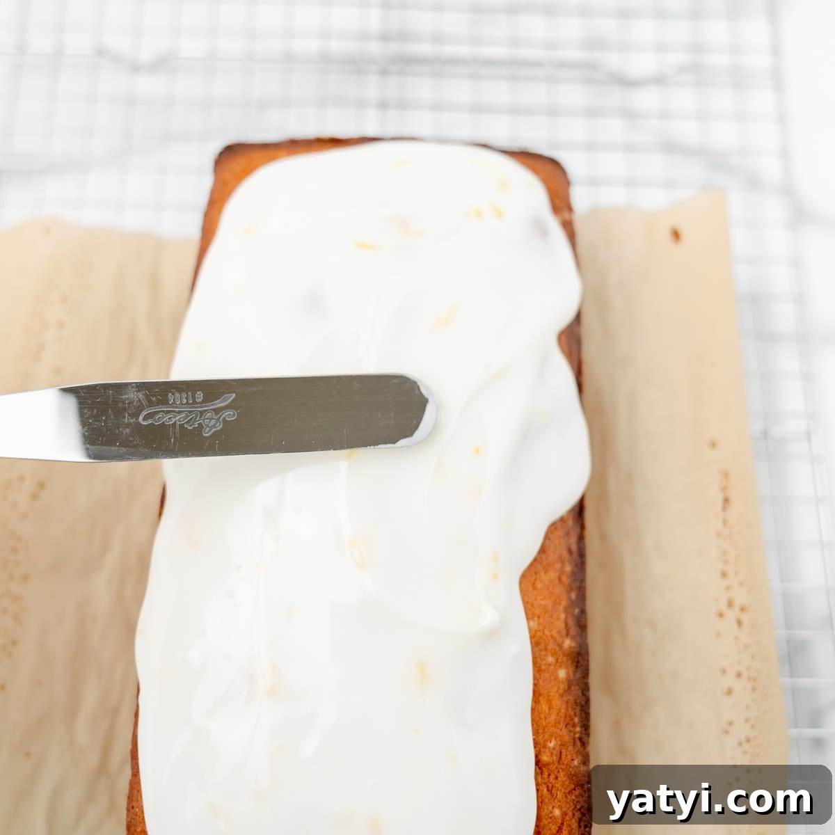 Copycat Starbucks Lemon Loaf Recipe: Moist Citrus Pound Cake Tutorial 9 A metal spatula spreads white icing over a rectangular loaf cake on parchment paper, set on a wire cooling rack.