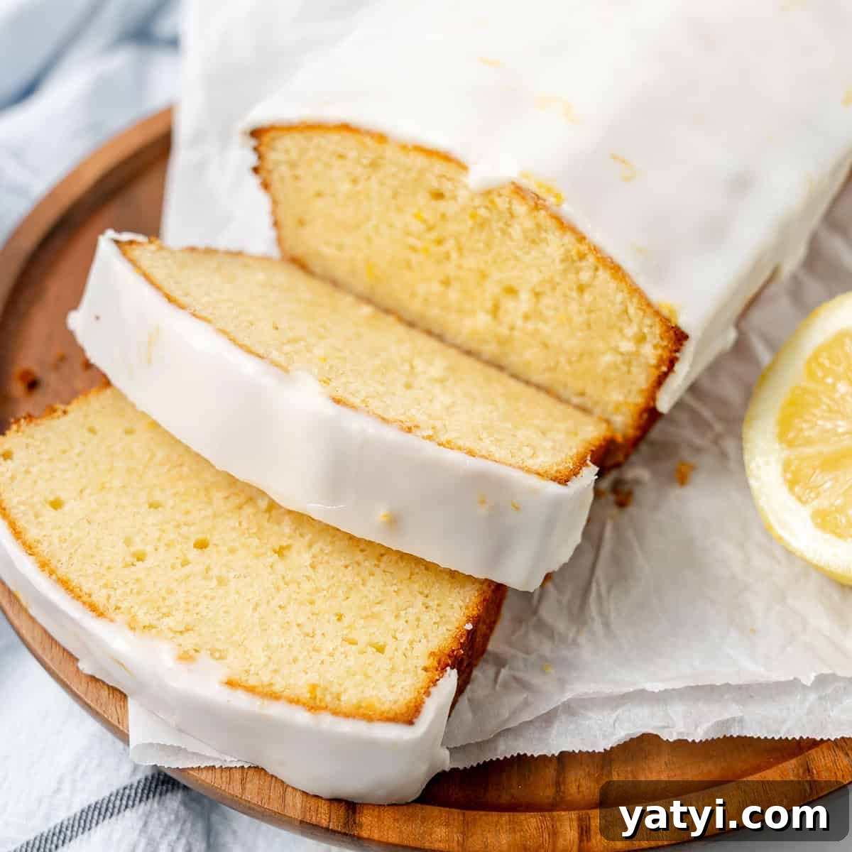 Copycat Starbucks Lemon Loaf Recipe: Moist Citrus Pound Cake Tutorial 10 A sliced loaf of lemon pound cake with white icing sits on a wooden plate, next to a slice of lemon.