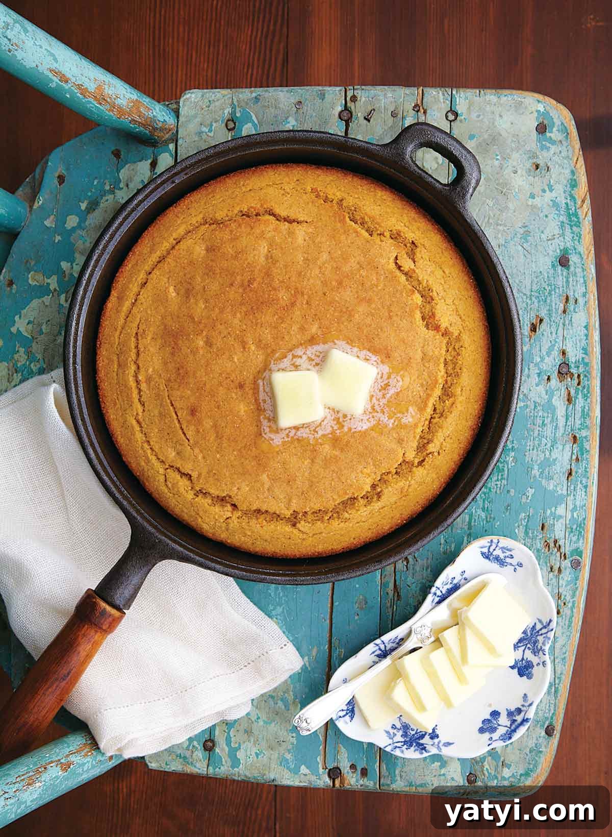 Sweet Potato Cornbread Recipe with Maple Butter Glaze 2 A cast-iron skillet filled with a cooked sweet potato cornbread with two pats of butter on top and a dish of butter pats beside it.