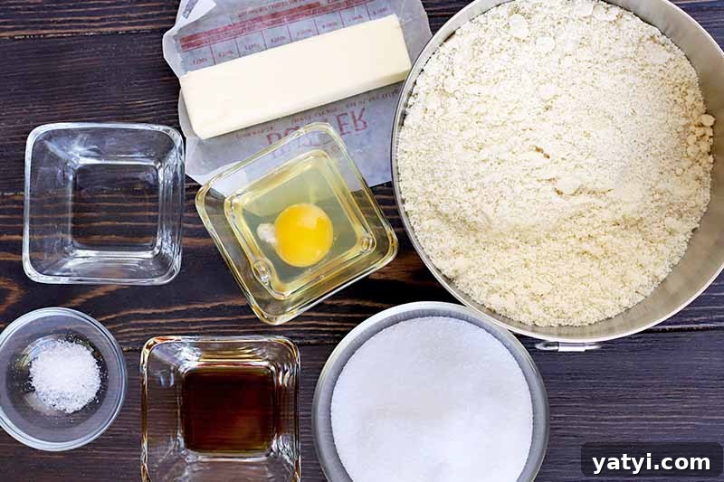 Overhead shot of a large bowl of almond meal with smaller round and square bowls of egg, sugar, vanilla extract, salt, and a stick of butter on a red and white waxed paper wrapper, on a dark brown wood surface.