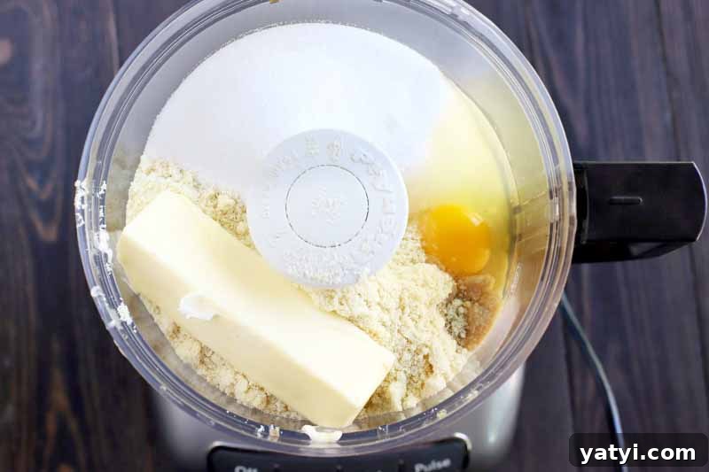A stick of butter, sugar, almond meal, and an egg in a plastic food processor canister, on a dark brown wood table.