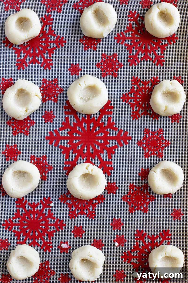 Top-down shot of cookie dough rolled into balls and pressed with a thumbprint indentation in the center of each, arranged in rows on a silicone pan liner with a red snowflake pattern.