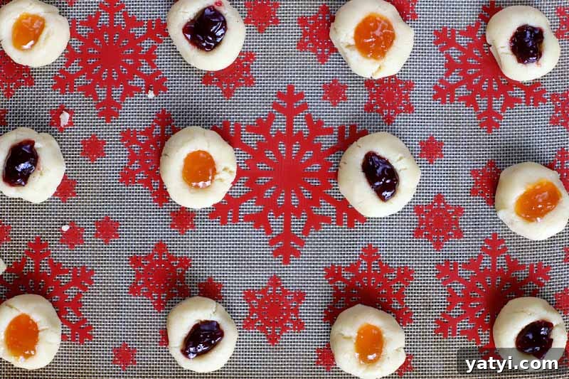 Over head shot of twelve almond thumbprint cookies filled with two types of fruit jam, arranged in three rows on a silicone pan liner decorated with a red snowflake pattern.