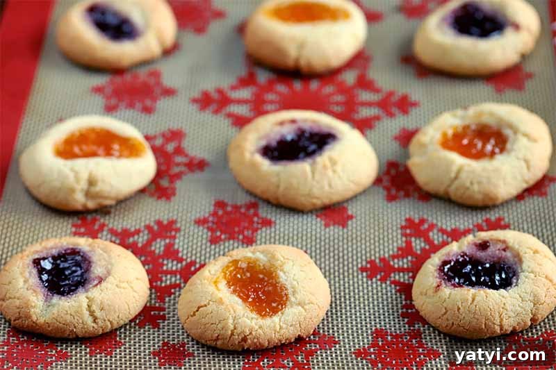 Oblique shot of thumbprint cookies filled with orange and purple-colored jam arranged on a red snowflake-patterned silicone baking sheet liner.
